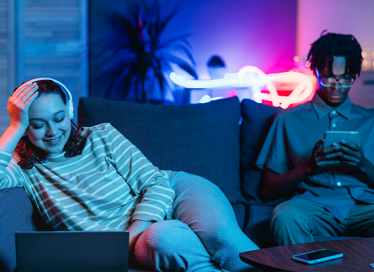 Image of two people on a couch; one smiling at a laptop with headphones, the other using a tablet.
